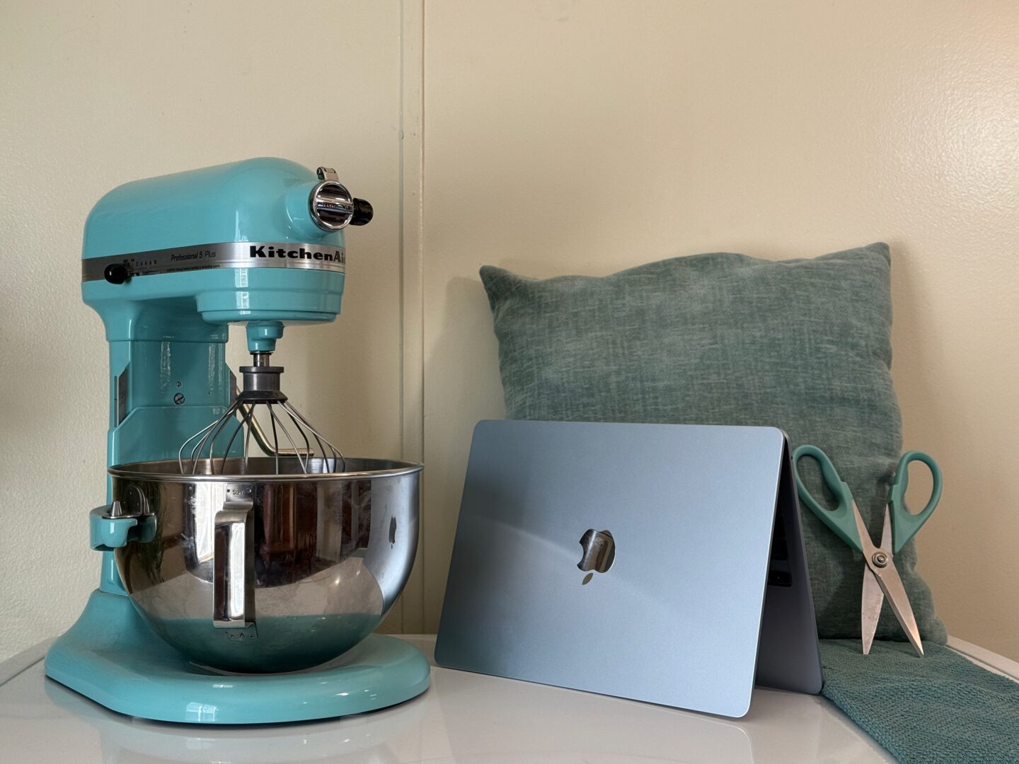 Sky blue M4 MacBook Air next to an aqua sky KitchenAid stand mixer, light blue pillow, scissors and towel